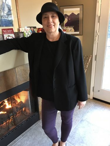 image of Casey Hobbs in a dark blazer, skinny jeans, and cute hat standing in front of her fireplace