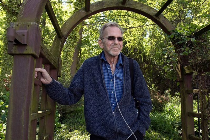 an image of an older man who has cancer standing outdoors wearing a blue sweater and dark sunglasses with his oxygen tank and line around his ears and nose