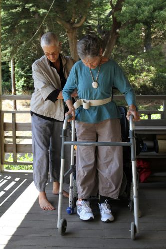 an outdoor image of two women, one is helping the other with her walker as she attempts to take some steps